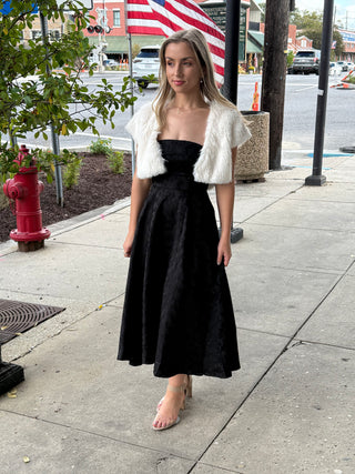 Woman in a black dress with a cream fuzzy vest standing on a sidewalk with an American flag in the background.