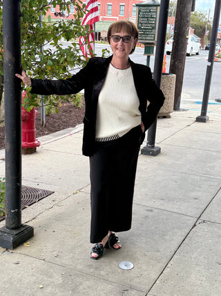 Woman in black blazer and pants standing on a sidewalk with a street and American flag in the background.
