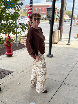 Woman standing on a sidewalk wearing sunglasses and a brown top with patterned pants.
