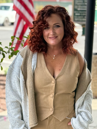 Woman with red hair wearing a beige vest and gray cardigan outdoors with an American flag in the background.