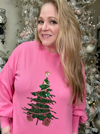 Woman wearing a pink sweater with a Christmas tree design in front of decorated trees.