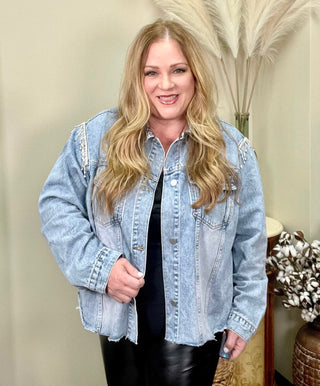 A woman wearing a cropped denim jacket with bling shoulder detail, standing in a room with a plant and furniture in the background.