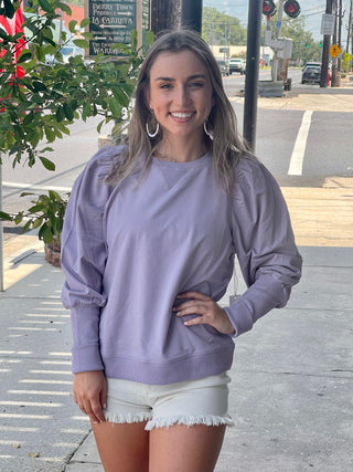 Woman standing on sidewalk with hand on her hip wearing a purple long sleeve sweatshirt