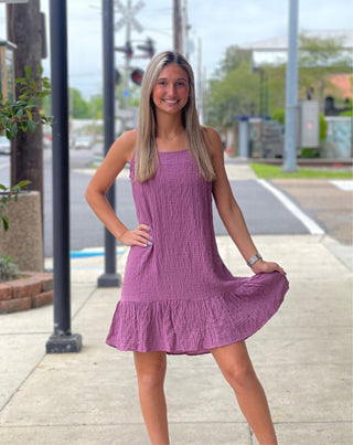 A woman standing on a sidewalk wearing a lavender-colored, short-sleeved, spaghetti strap, ruffle skirt dress.