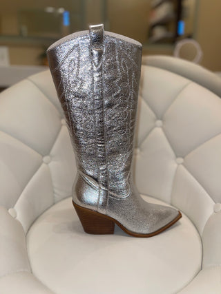A silver metallic boot with a high heel and pointed toe, displayed on a white chair.