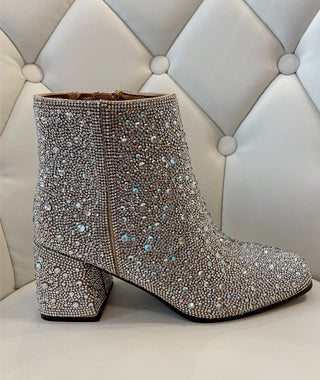 A pair of sparkling clear rhinestone-studded boots on a white surface with a white, quilted bench in the background.
