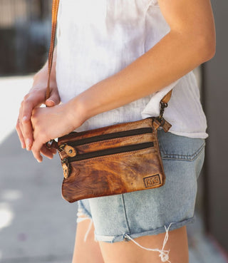 A person wearing a white top and blue shorts is carrying a brown, solid-colored, crossbody purse with a zipper.
