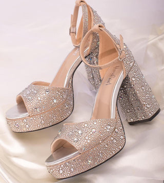 A pair of clear rhinestone-embellished heels with a sandal style, featuring a shiny metallic finish and a label with 'HeyGirl by Corkys' on the inside of the shoe on the heel