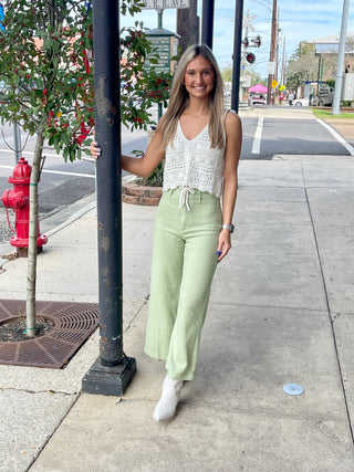 A woman standing on a sidewalk next to a street column, wearing green cropped wide-leg pants with front pockets and a white crotchet top with off white boots.