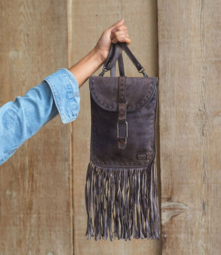 A person holding a brown BedStu Sandy Lane Alkaline DD handbag with fringe detail against a wooden background.