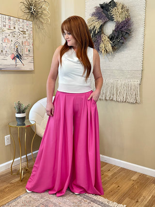 A woman standing with wide leg magenta pants on paired with a white silk top.