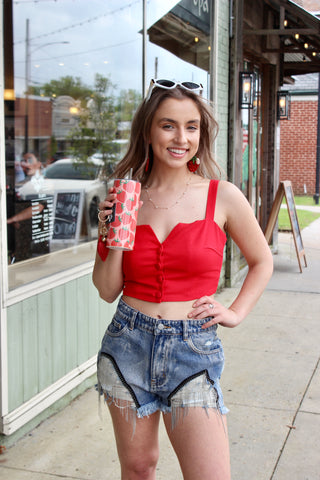 A person wearing red denim shorts with a metal fringe design, holding a strawberry print cup, with a storefront in the background.