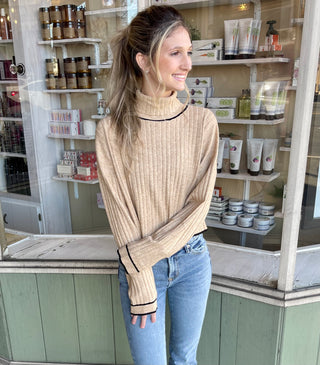Woman wearing a oatmeal ribbed long sleeve turtleneck with black accents, paired with blue denim jeans