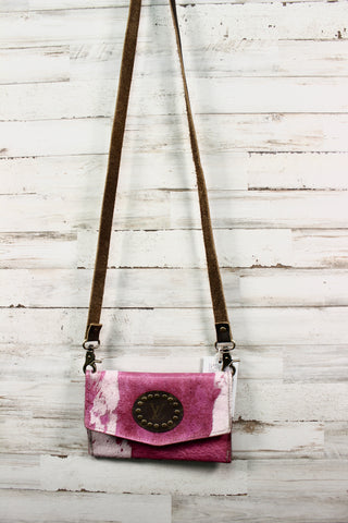 Upcycled cow print purse with light pink and magenta colors, with a brown leather strap hanging in front of modern white wood backdrop.