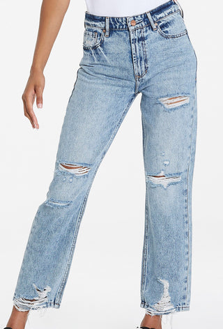 Woman standing in front of white background wearing light distress denim jeans.
