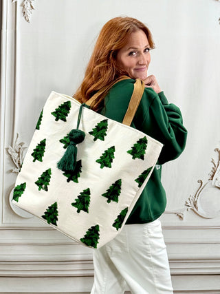 A woman with red hair wearing a green sweatshirt and white pants carries a large off-white canvas tote bag decorated with green embroidered Christmas trees and a green tassel drawstring, set against a white paneled wall.
