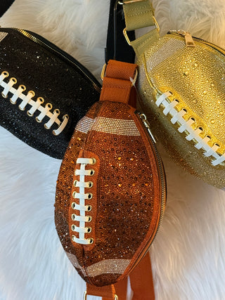 Three football-shaped crossbody bags with a glittery finish, arranged on a white faux fur background. The center bag is copper-brown glitter with white laces, flanked by a black glitter bag on the left and a gold glitter bag on the right.