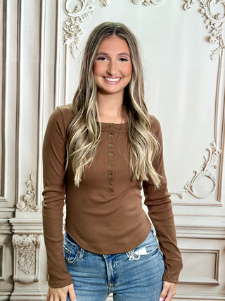 A smiling woman with blonde-highlighted hair wearing a brown long-sleeve henley top and blue jeans, standing in front of an ornate cream-colored wall.