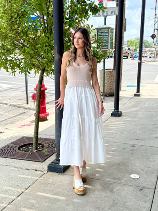 Woman in a dress that has beige top and white skirt standing on a sidewalk with a street and trees in the background.
