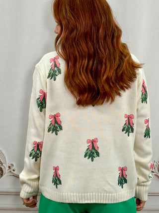 A woman viewed from behind wearing a cream-colored knit sweater with green holly leaves and pink bows, paired with green pants.