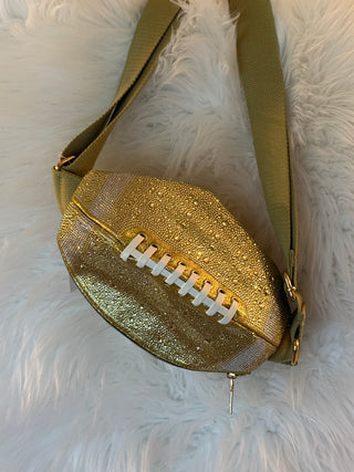 A football-shaped shoulder bag with a glittery gold body, white laces, and an olive green strap, displayed on a white faux fur background.