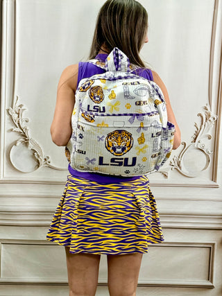 A girl seen from behind wearing a purple and yellow LSU tiger-themed backpack, a purple top, and a matching purple and yellow patterned pleated skirt, standing in front of an ornate white wall.