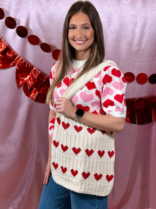 Woman wearing a heart-patterned sweater holding a matching bag with red and pink hearts on a pink background.