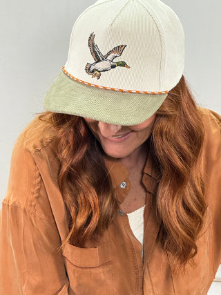 A woman wearing a cream-colored ribbed baseball cap with a light green visor and an embroidered mallard duck on the front, paired with a tan overshirt.
