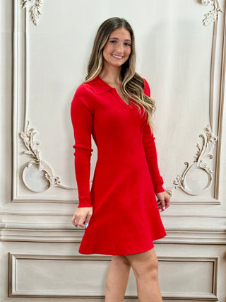 A woman modeling a bright red long-sleeve mini dress with a collared neckline, standing against an ornate beige wall.