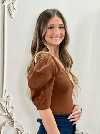 A woman with long blonde-highlighted hair wearing a brown puff-sleeve top and blue jeans, posing in front of a white decorative wall.