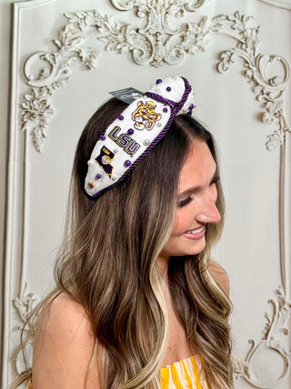 A woman with long brown and blonde highlighted hair wears a white LSU Tigers themed headband with purple trim and embroidered details, posed in front of an ornate white wall.