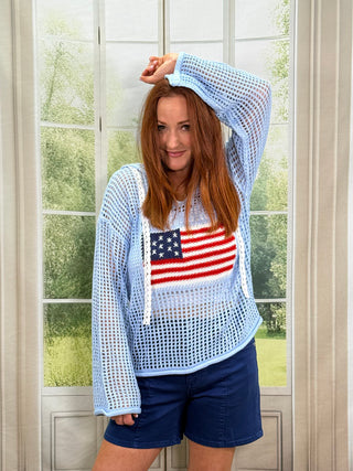 Woman wearing a light blue mesh top with an American flag design and dark blue shorts, standing indoors.
