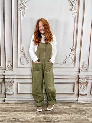 A woman with red hair stands in front of an ornate gray wall, wearing olive green corduroy bib overalls over a white long-sleeve shirt and brown sneakers.
