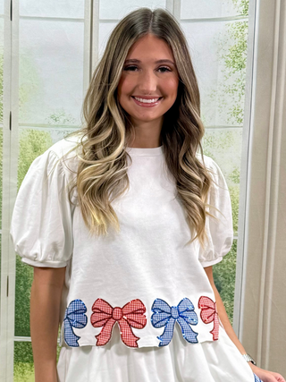 Woman wearing a white blouse with red and blue bow designs in front of a window with greenery outside.
