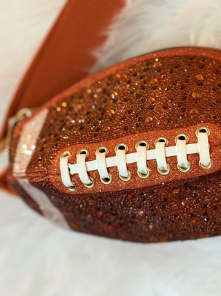 A close-up of a football-shaped clutch bag covered in coppery brown glitter, with faux white laces and gold eyelets, resting on a white furry surface.