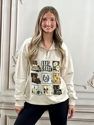 A woman with long blonde hair wearing a cream-colored quarter-zip sweatshirt with a graphic collage print on the front, paired with black pants, standing in front of a white paneled wall.
