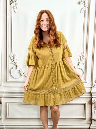 A woman with long red hair wearing a moss cotton dress with lace flutter sleeves and a button-up front, posing in front of a white ornate wall.