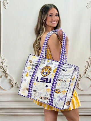 A woman wearing a yellow and white striped dress holds up a large LSU Tigers themed tote bag. The tote bag has a white quilted body with purple gingham trim, featuring the LSU logo, tiger mascot, and other team graphics.