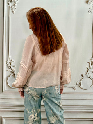 A woman with long auburn hair is shown from behind, wearing a light blush pink sheer long-sleeve blouse and light blue floral print pants, standing against an ornate white wall.