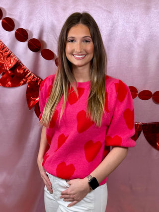 Woman wearing a pink sweater with red heart patterns against a decorative background.