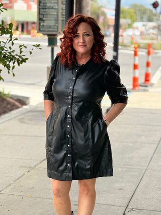 Woman wearing a black leather dress standing on a sidewalk with a city street background.