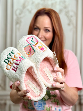 A woman holds a pair of fluffy off-white slippers with colorful embroidered patches and text. The slippers have a pink lining and a visible brand tag reading 'FELINE FASHION BRAND'.