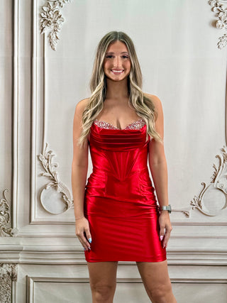 A woman with blonde hair wearing a shiny red strapless cocktail dress with beaded embellishments on the sweetheart neckline, standing in front of a white ornate wall.