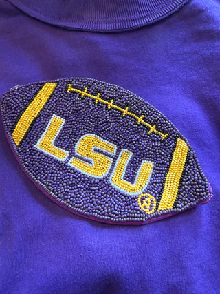 A close-up of a purple beaded patch shaped like a football on a matching purple garment. The patch features the letters 'LSU' in yellow with white and purple outlines, yellow lacing details, and yellow side stripes.