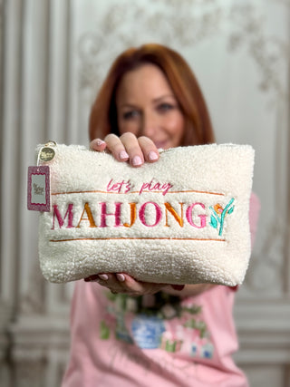 A woman holds up a cream-colored plush fleece zippered pouch with colorful embroidered text reading 'let's play MAHJONG' and a small green leaf motif. The background is a soft-focus indoor setting with light-colored columns.