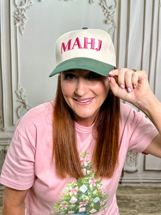 A woman wearing a cream and sage green baseball cap with 'MAHJ' embroidered in pink on the front, paired with a pink t-shirt featuring a Christmas tree graphic, posed in front of an ornate white paneled wall.