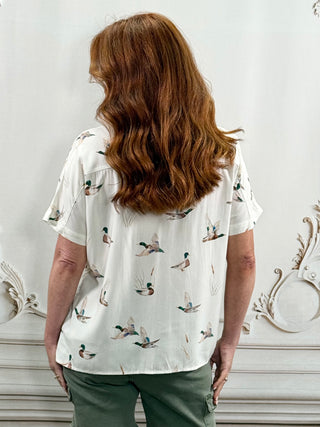 A woman with long wavy auburn hair is shown from behind, wearing a short-sleeve white button-up shirt with a scattered duck print and olive green pants, standing in front of a white wall with decorative molding.