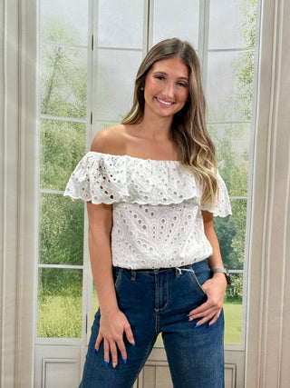 Woman wearing a white off-shoulder top with floral patterns and blue jeans, standing in front of a window with greenery outside.