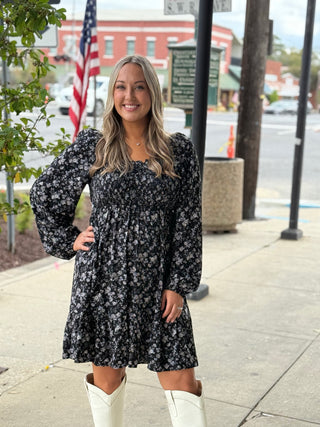 Woman in a floral dress and white boots standing on a sidewalk with an American flag in the background.