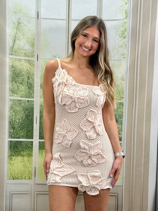 Woman wearing a beige crochet dress standing in front of a glass door with greenery outside.